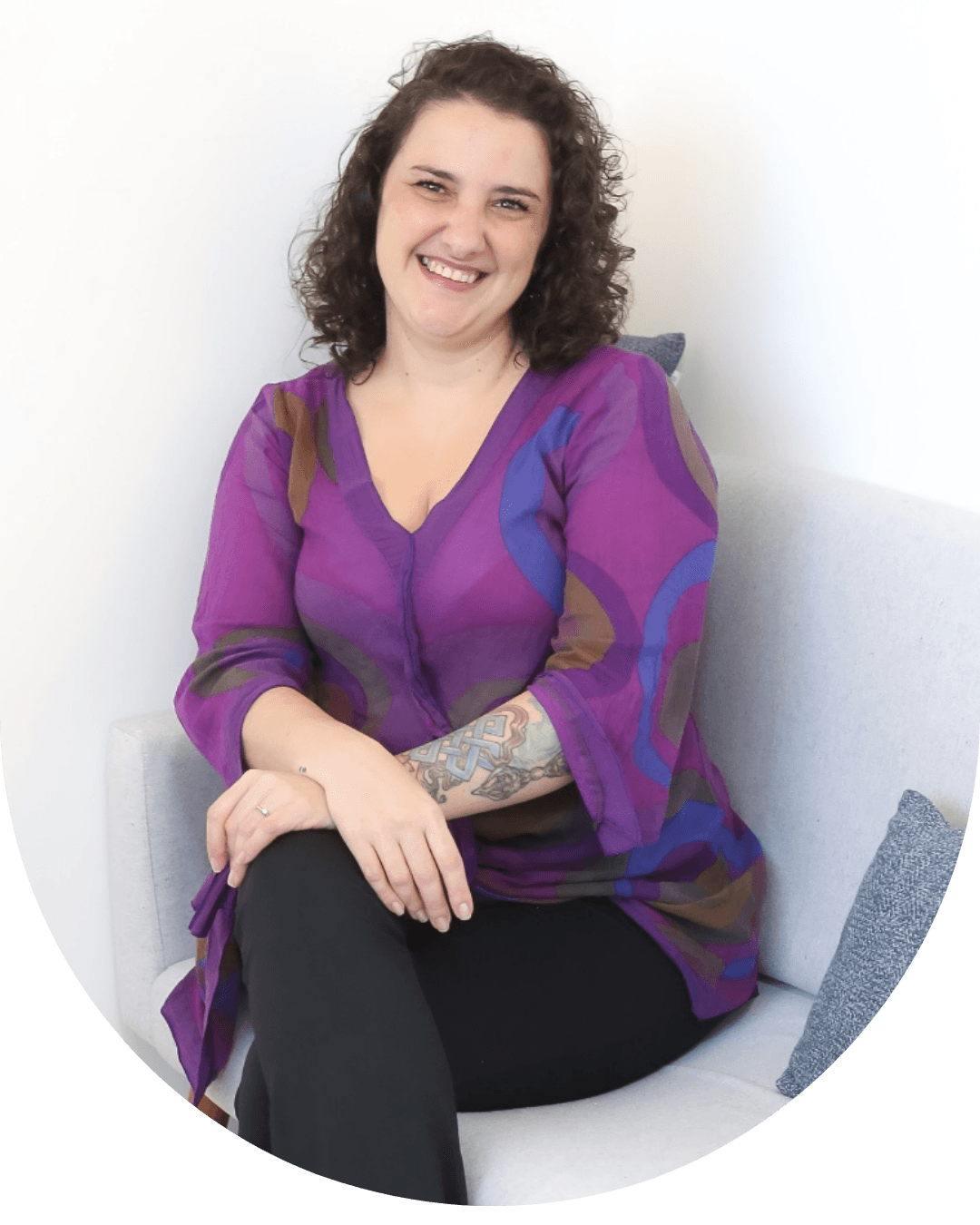 Psicóloga e neuropsicóloga Audrey Ruggiero Pilli sentada em uma poltrona clara, sorrindo para a câmera, usando blusa roxa estampada e calça preta, em um ambiente iluminado.